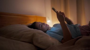 Yatakta uzanan genç bir kadın, yüzünde hafif bir lamba, ışık saçan akıllı telefon özellikleri.,