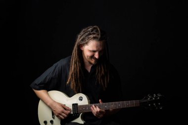 Elinde elektro gitar tutan, gözleri kapalı, gülümseyen, siyah bir arka plana bakan uzun saçlı bir adam.