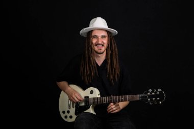Bir müzisyen elektro gitar çalıyor, beyaz şapka ve siyah gömlek giyiyor, uzun rastalı, siyah arka planda