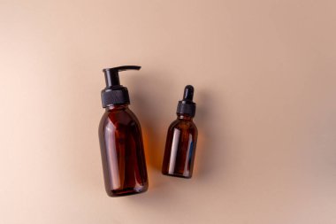 Kehribar cam şişeleri, birinde pompa dağıtıcısı, diğerinde de damlatıcı var, düz, açık renkli bir yüzeye yerleştirilmiş, cilt bakımı ya da aromaterapi ürünleri öneriliyor.