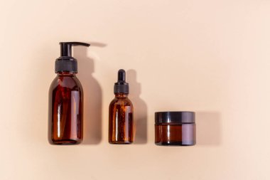 Bir pompa şişesi, bir damlatıcı şişesi ve bir kavanoz da dahil olmak üzere üç kehribar cam kap, yumuşak gölgeli nötr bir arka planda düzenlenmiştir.