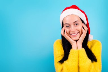 Güzel bayan kollarının fotoğrafı Noel Baba şapkasıyla izole edilmiş mavi perde arkasında elmacık kemiklerine dokunuyor.