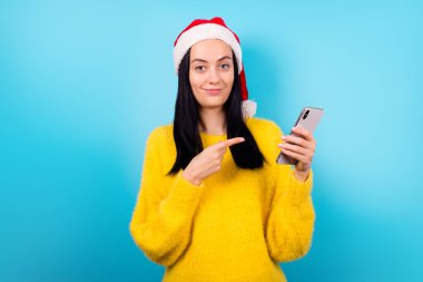Heyecanlı komik bayanın fotoğrafı. Noel Baba şapkası, telefonun mavi renk arka grubunda izole olduğunu gösteriyor.