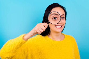Sevimli, neşeli genç bir kadının fotoğrafı göz doktorunu ziyaret ediyor mavi renk arka grubunda izole edilmiş büyüteçle gözlerini kontrol ediyor.