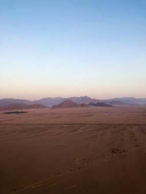Bir hava manzarası, eğimli bir gökyüzü altında, engebeli dağları ve uzak bölgeleri birbirine bağlayan ıssız bir yolu gösteren devasa bir çöl ortamı ölçer.