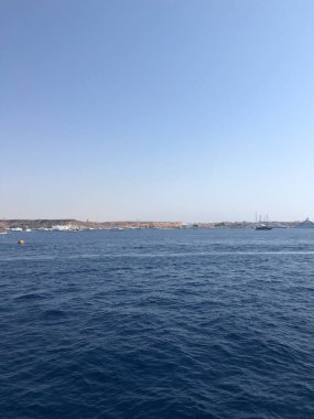 Derin mavi denizin uçsuz bucaksız genişliği, binaları ve tekneleri olan bir sahil şehrinin parlak, bulutsuz bir gökyüzünün altında görülebildiği puslu bir ufka doğru uzanıyor.