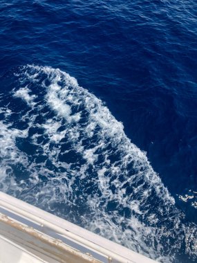 Engin, derin mavi denizde ilerleyen bir geminin arkasında sallanan çalkantılı beyaz dalganın yakın görüntüsü.