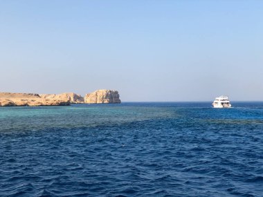 Beyaz bir yolcu gemisi derin mavi denizde, kayalık, kurak bir kıyı şeridinde yüzeysel turkuaz sularda yüzer ve aşağıdaki mercan resiflerini gösterir.