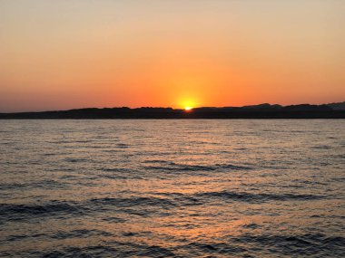 Güneş ufkun altına batıyor, sakin denizi ve uzak tepeleri sıcak bir şekilde aydınlatıyor.