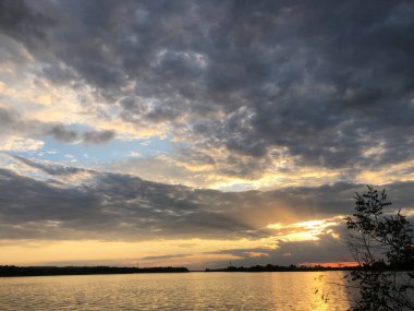 Güneş, sakin bir göl boyunca altın ışık saçan ağaçların arkasında batar dramatik bulutlar tepemizde toplanır.