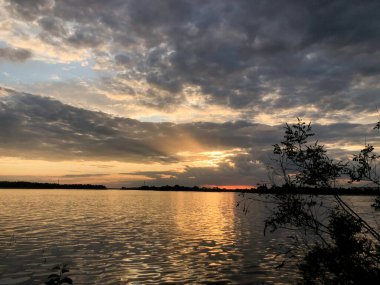 Altın saat ışığı dalgalanan suya yansıyor güneş kasvetli bir gökyüzünün altında uzak bir ağaç hattının ardında batarken.