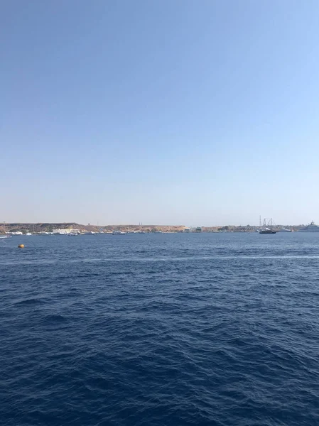 Derin mavi denizin uçsuz bucaksız genişliği, binaları ve tekneleri olan bir sahil şehrinin parlak, bulutsuz bir gökyüzünün altında görülebildiği puslu bir ufka doğru uzanıyor.