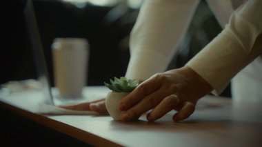 Bir insanın elleri, arka planda dizüstü bilgisayarı ve içkisi olan küçük bir saksıyı bir masaya yerleştirirken görülüyor.