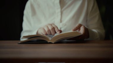 Beyaz gömlek giyen bir kişi, bir kitap okumaya dalmış, parmakları yavaşça koyu ahşap bir yüzeyde sayfaları çeviriyor.