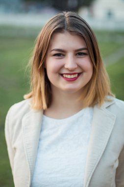 Sarışın ve kırmızı rujlu neşeli genç bir kadın açık havada açık renkli bir ceket ve beyaz bir bluz giyerken sıcacık gülümsüyor.