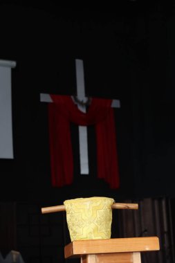 Dikenli bir taç ve kırmızı kumaşla süslenmiş, ahşap sunaktaki altın bir kadehin üzerine yerleştirilmiş beyaz bir haçın yer aldığı kutsal bir dini sahne.