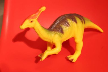 Sarı ve kahverengi plastik bir Parasaurolophus dinozor oyuncağının detaylı görüntüsü kırmızı bir yüzeye gölge düşürüyor.