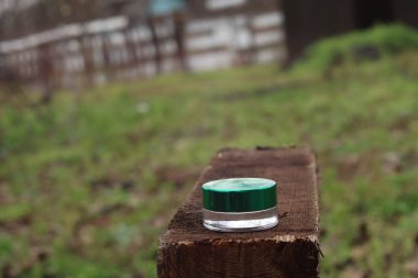 Metalik yeşil kapaklı tek bir cam kavanoz, bulanık yeşillik ve kırsal bir zemin ile çevrili, kaba bir tahta üzerinde oturmaktadır.