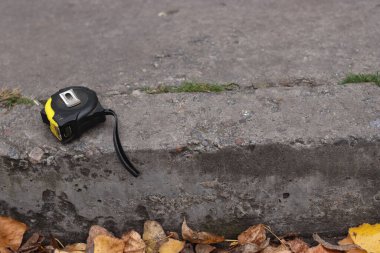 Dayanıklı bir mezura, kurumuş yapraklar ve çimlerle çevrili eskimiş beton kaldırımın kenarında yatıyor.