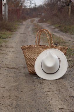 Kuru bitki örtüsüyle çevrili tozlu bir yola şık bir hasır çanta ve klasik bir fötr şapka yerleştirilir.