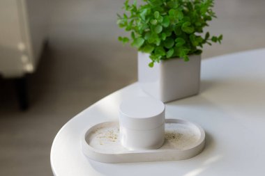 Temiz ve basit bir kompozisyon, beyaz bir kozmetik kavanozu dekoratif bir tepside, parlak beyaz bir yüzeyde, küçük yeşil bir saksı bitkisinin yanında.