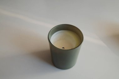 Beyaz balmumu ve koyu fitili olan yeşil cam bir kavanozun yakın plan çekimi, düz bir arkaplanda ince bir gölge bırakır.