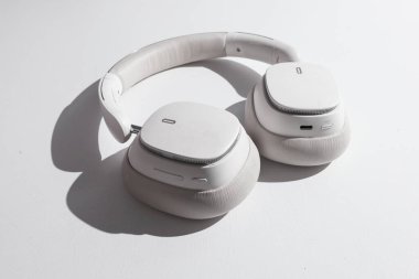 Bir çift parlak, çağdaş beyaz kulaklık, çarpıcı stüdyo ışıklandırması ve belirgin bir gölgeyle vurgulanmış, desenli beyaz bir yüzeye dayanır.