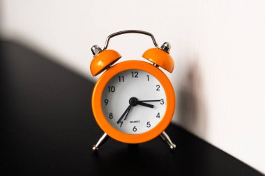 Parlak turuncu bir ikiz çan çalar saati siyah bir yüzeye oturur, beyaz bir duvara gölge düşürür, klasik tasarımını ve net zaman göstergesini vurgular.