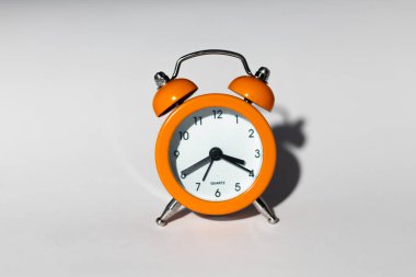 Beyaz yüzlü, siyah elli, klasik turuncu alarm saati beyaz bir yüzeye oturur ve ince bir gölge bırakır.