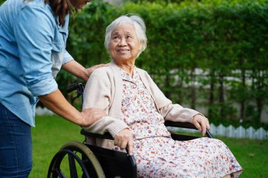 Bakıcı, parkta tekerlekli sandalyede oturan Asyalı yaşlı kadın maluliyet hastasına yardım ediyor..