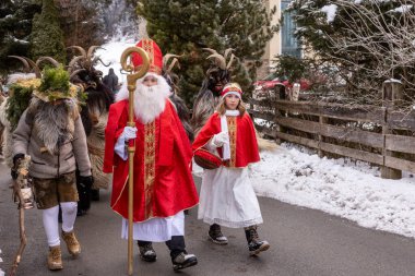Kış kostümlü birkaç kişi bir köyde karlı bir yolda yürüyor. Bir figür kırmızı bir cübbe giyer ve bir asa tutarken, diğerleri hayvan maskeleri takar. Kar zemini kaplıyor..