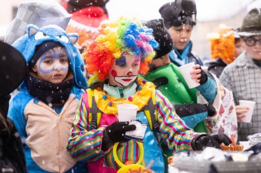 Palyaço gibi giyinmiş bir çocuk, bir kış festivalinde elinde bir kupa tutarken etrafta kar yağıyor.