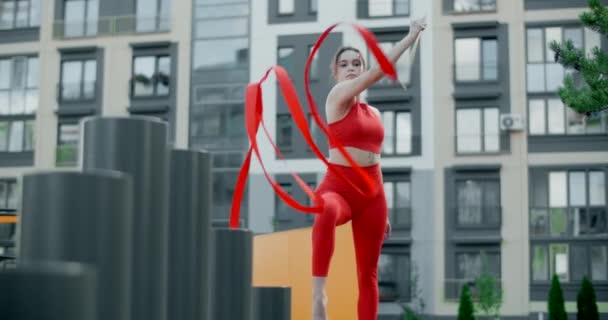 Jeune femme en rouge effectue callisthenics avec ruban dans la cour de libing appartement, gymnaste fait des exercices acrobatiques dans une zone résidentielle, fitness dans l'environnement urbain, 4k 120p Prores HQ — Video