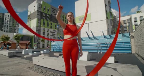 Mujer joven en rojo realiza calistenia con cinta en el patio de entrenamiento al aire libre, gimnasta hace ejercicios acrobáticos en un área residencial, aptitud en el entorno urbano, 4k 120p HQ de Prores — Vídeo de stock