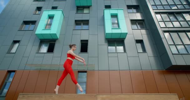 Mujer gimnástica en rojo hace ejercicios acrobáticos en el banco en el patio de un edificio de apartamentos, fitness en el entorno urbano, calistenia al aire libre, entrenamiento físico, 4k 120p Prores — Vídeos de Stock