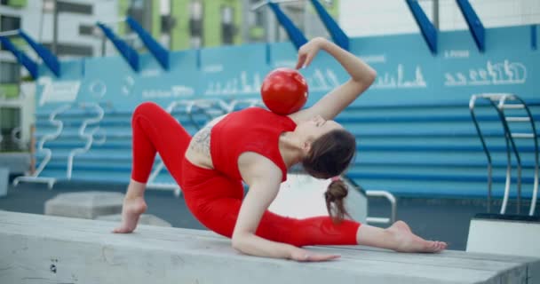 Žena v červeném sportovním kostýmu provádějící kallisthenická cvičení s gymnastickým míčkem na cvičišti venku, sport v městském prostředí, gymnastika ve městě, 4k 120p Prores HQ — Stock video