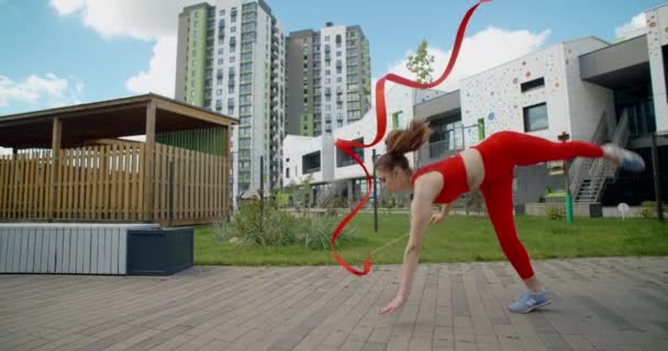 Ung atletisk kvinde i rødt udfører vejrmølle med gymnastikbånd i gården af en lejlighedskompleks, gymnastik i byen, træning udendørs, sport i slowmotion, 4k 120p Prores HQ 10 bit – Stock-video