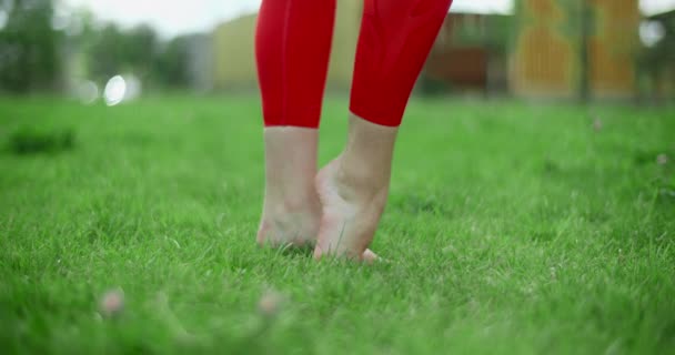 Genç bayan jimnastikçi, yeşil çimenler üzerinde antrenman yapmadan önce bacağını ısıtıyor, şehir ortamında spor yapıyor, yoga sınıfında açık havada, şehirde fiziksel antrenman yapıyor, 4k 120p Prores — Stok video