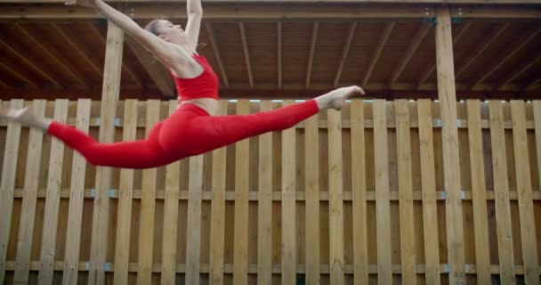 Jeune gymnaste femme en rouge saute au ralenti, femme effectue grand jete en plein air, acrobaties dans la ville, l'entraînement en plein air, callisthenics au ralenti, 4k 120p Prores HQ 10 bits — Video