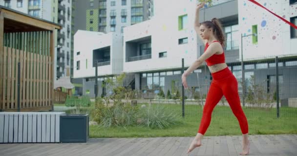 Mujer atlética joven en rojo realiza voltereta con cinta de gimnasia en el patio de un edificio de apartamentos, gimnasia en la ciudad, entrenamiento al aire libre, deportes en cámara lenta, 4k 120p HQ Prores 10 bit — Vídeo de stock