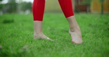 Genç bayan jimnastikçi, yeşil çimenler üzerinde antrenman yapmadan önce bacağını ısıtıyor, şehir ortamında spor yapıyor, yoga sınıfında açık havada, şehirde fiziksel antrenman yapıyor, 4k 120p Prores