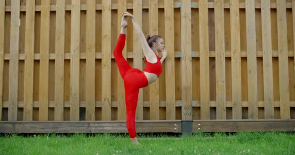 Woman in red does stretching and yoga exercices for legs in the yard of an apartment building, fitness in the urban environment, yoga class outdoors, physical workout in the city, 4k 120p Prores — Stock Video