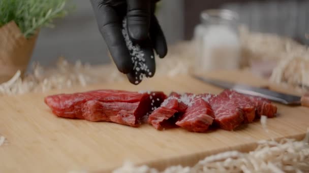Metzger in schwarzen Handschuhen gibt etwas Salz zu den rohen Laiben in Zeitlupe, bereitet den Grill zu, kocht Fleisch, Full HD 240 fps Prores HQ — Stockvideo
