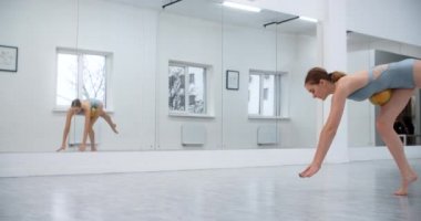 Kadın sporcu, beyaz dans salonunun zemininde jimnastik topuyla spor egzersizleri yapıyor, ayna duvarının önünde spor egzersizleri, beyaz salonda jimnastik, 4k 120p Prores HQ