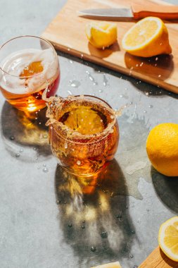 Gri arka planda limonların yanında su sıçratan bir bardağın yakın plan görüntüsü.