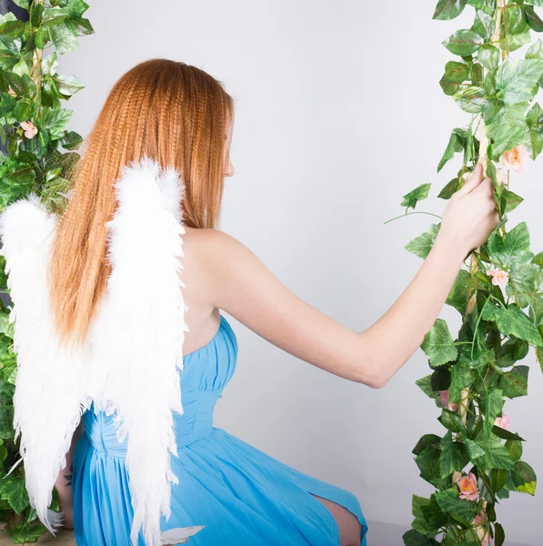 Beautiful young leggy redhaired woman in a long blue dress on a swing, wooden swing suspended from a rope hemp, rope wrapped vine and ivy. angel wings behind — Stock fotografie