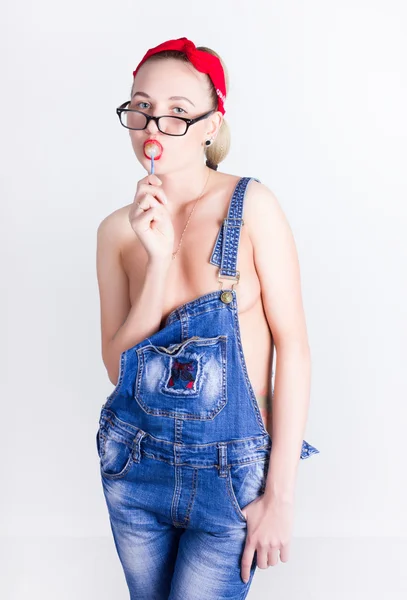 Menina loira bonita e jovem em uma bandana vermelha, macacão jeans sobre seu corpo nu, lambendo doces — Fotografia de Stock