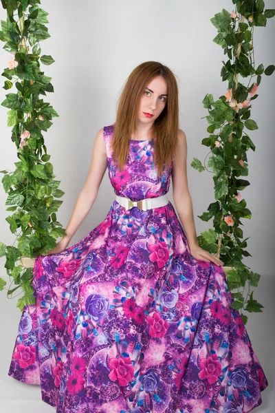 Beautiful young leggy redhaired woman in a long colorful dress on a swing, wooden swing suspended from a rope hemp, rope wrapped vine and ivy — Stock Photo, Image