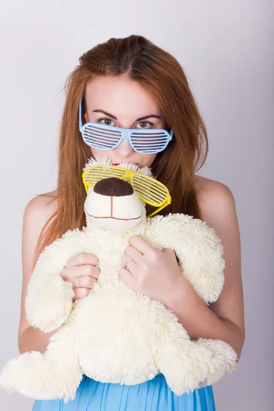 Girl in a blue dress and sunglasses in the style of disco, hugging a teddy bear in the same glasses — Stock fotografie