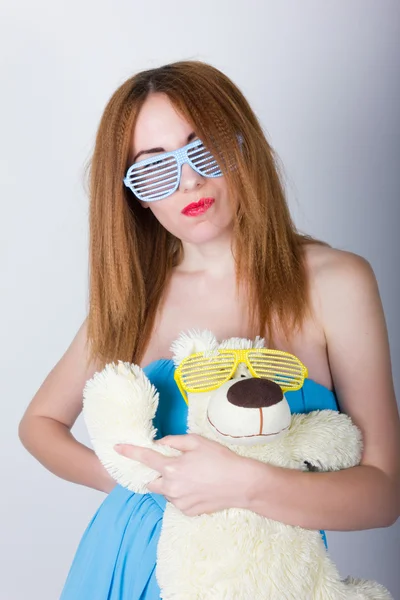 Chica en un vestido azul y gafas de sol en el estilo de discoteca, abrazando a un oso de peluche en los mismos vasos — Foto de Stock
